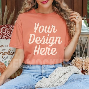 May include: A woman wearing a peach colored t-shirt with the text "Your Design Here" printed on the front. She is sitting on a couch with a white throw blanket and fall decor.