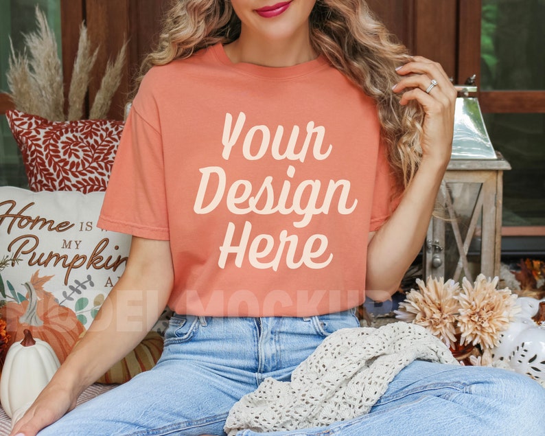 May include: A woman wearing a peach colored t-shirt with the text "Your Design Here" printed on the front. She is sitting on a couch with a white throw blanket and fall decor.