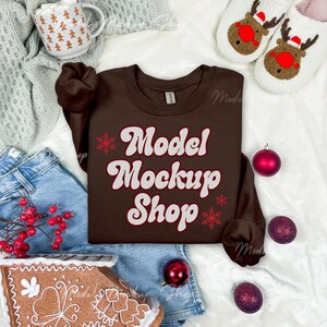 May include: A brown sweatshirt with the text "Model Mockup Shop" in white with red accents. The sweatshirt is laid out on a white surface with a gingerbread house, blue jeans, and red and purple ornaments.