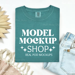 May include: A teal blue t-shirt with the text "MODEL MOCKUP SHOP" in white. The text is in a retro font style with stars on either side. The text "REAL POD MOCKUPS" is below the main text.