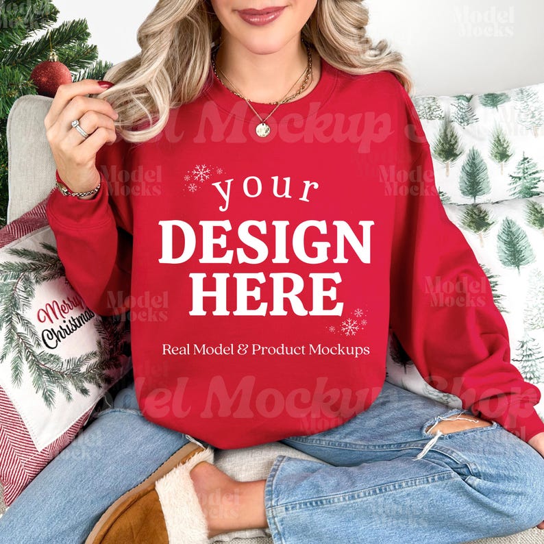 May include: A red crewneck sweatshirt with the text "your DESIGN HERE" in white. The sweatshirt is part of a product mockup, with a Christmas theme. The model is wearing blue jeans and sitting on a couch with Christmas pillows.
