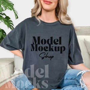 May include: Dark gray t-shirt with the text "Model Mockup Shop" in a bold, black font. The shirt has a relaxed fit and is worn with blue denim shorts. The overall aesthetic is casual and comfortable, suitable for showcasing designs.