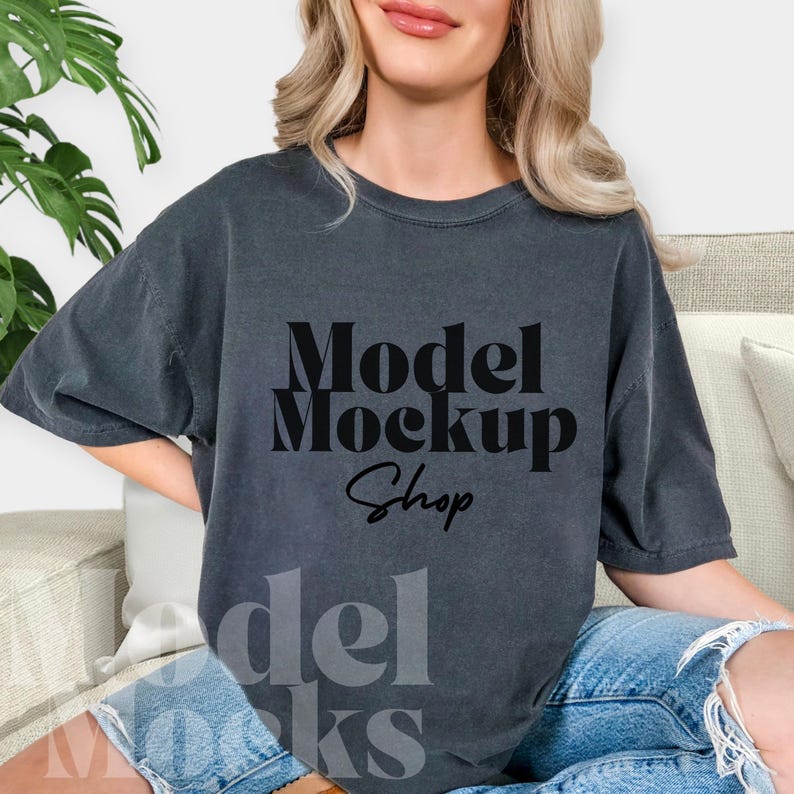 May include: Dark gray t-shirt with the text "Model Mockup Shop" in a bold, black font. The shirt has a relaxed fit and is worn with blue denim shorts. The overall aesthetic is casual and comfortable, suitable for showcasing designs.