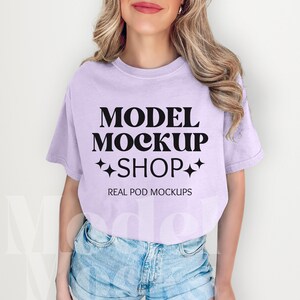 以下が含まれることがあります： モデルは、ラベンダー色のTシャツを着ています。Tシャツには「MODEL MOCKUP SHOP」と「REAL POD MOCKUPS」の文字が印刷されています。モデルはデニムのショートパンツを履いています。