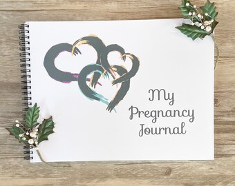 May include: A spiral-bound journal with the text "My Pregnancy Journal" on the cover. The cover features three overlapping heart shapes in shades of gray, teal, purple, and peach. Decorative holly and berries are in the corners.