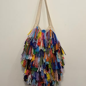 May include: A handmade, colorful tote bag with a cream-colored strap and a dense, shaggy fringe. The fringe is composed of various yarn colors, including blue, green, yellow, orange, and purple. The bag is suspended against a white wall.