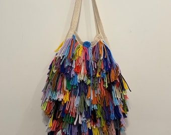 May include: A vibrant, handmade tote bag featuring a cream-coloured strap and a dense, shaggy fringe. The fringe is made from various yarn colours, including blue, green, yellow, orange, and purple. The bag hangs against a white wall.