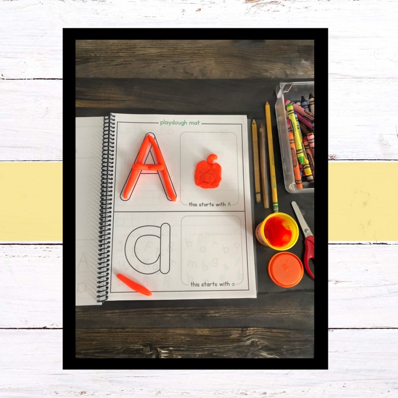 May include: A playdough mat with the letter A and an apple made out of playdough. The mat also has a section for tracing the letter a. There are crayons, pencils, playdough, and scissors on the table.