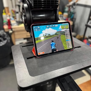 May include: An exercise bike with a tablet showing a cycling simulation. The tablet, in a red case, is mounted on a black platform. A black fan is positioned above the tablet, enhancing the indoor cycling experience.