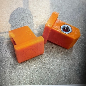 Puede incluir: Dos conectores de plástico naranja impresos en 3D con un agujero roscado. Los conectores tienen una base cuadrada y una parte superior rectangular.