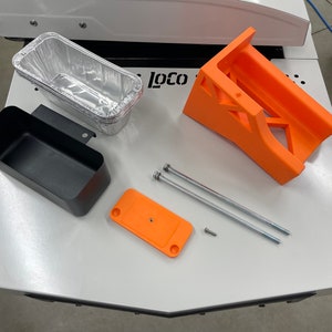 May include: A set of 3D printed parts for a 3D printer, including a black storage bin, an orange bracket, an orange plate, and two silver metal rods with nuts. The parts are on a white surface with the word "loco" printed on it.