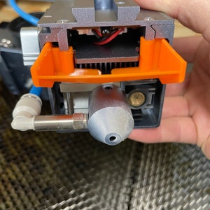 Puede incluir: Primer plano de una boquilla de impresión 3D con una carcasa de plástico naranja. La boquilla está unida a un dispositivo metálico plateado y negro con un cable azul.