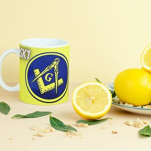 May include: A white ceramic mug with a yellow background and a blue circle containing a yellow and gold compass and square with the letter "G" in the center. The text "SK7" is visible on the mug. The mug is sitting on a light brown surface with lemons and green leaves.