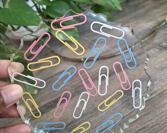 Puede incluir: Posavasos transparente de forma irregular con clips de colores. Los clips son de varios colores, incluyendo rosa, amarillo, azul y blanco. El posavasos está sostenido por una mano.
