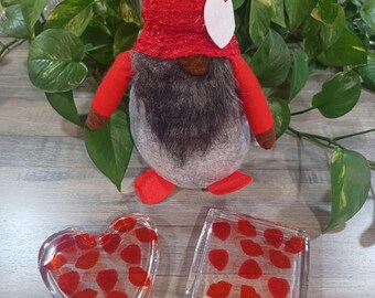 May include: Two clear glass coasters with red heart designs. One coaster is shaped like a heart and the other is square. A red gnome with a white heart on its hat is in the background.