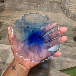 May include: A clear, round coaster with a wavy edge, featuring a vibrant blue ink design. The coaster is held in a hand, showcasing the color and design. The coaster is a decorative item for home decor.