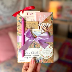 May include: A rectangular gift package wrapped in floral kraft paper, tied with a lavender ribbon, and topped with a red ribbon. It contains a TBR card, a pink tea packet, a pen, and cards with text including Jesus Christ the same yesterday.