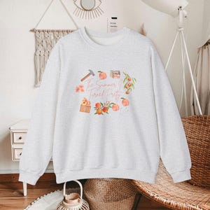 Peut inclure: Un sweat-shirt gris clair avec un motif de pêches, de fleurs et le texte "The Summer I Turned Pretty". Le motif comprend un marteau et une petite caisse en bois. Le sweat-shirt a des manches longues et un col rond.