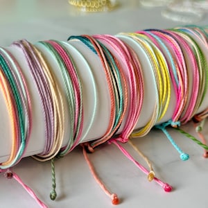 以下が含まれることがあります： ピンク、ブルー、グリーン、イエローなど、さまざまな色合いのカラフルな紐のブレスレットのコレクション。ブレスレットは一緒に結ばれており、扇形に配置されています。