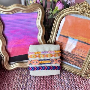 May include: A set of four colorful friendship bracelets with a variety of patterns, including chevron, diamond, and stripes. The bracelets are displayed on a white surface in front of two gold-framed pictures.