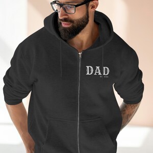 May include: A black zip-up hoodie with the word "DAD" printed in white on the chest. The hoodie has a drawstring hood and a full-length zipper. The text "EST. 2023" is below the word "DAD".