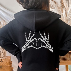 May include: Black hoodie with a white skeleton hand design making a heart shape with the word "WIFE" inside the heart.