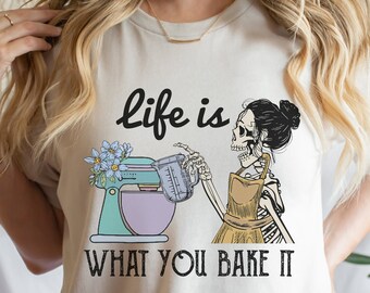 May include: A white t-shirt with a graphic of a skeleton holding a measuring cup and a stand mixer. The text "life is what you bake it" is printed below the graphic.