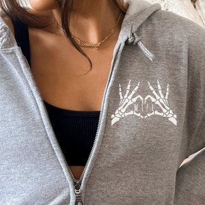 May include: Grey hooded sweatshirt with a white skeleton hand design that says "WIFE" in the centre of the heart.