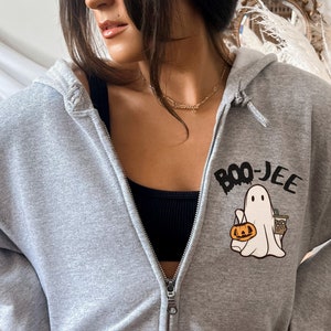 May include: A gray zip-up hoodie featuring a Halloween-themed graphic. The graphic includes a cartoon ghost holding a pumpkin and a coffee cup, with the text "BOO-JEE" above. The hoodie has a hood and a zipper closure.
