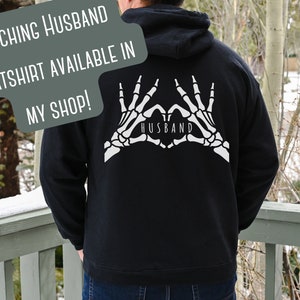 May include: Black hooded sweatshirt with a white skeleton hand design making a heart shape with the word "Husband" inside. The text "Matching Husband Sweatshirt Available in My Shop!" is superimposed on the image.