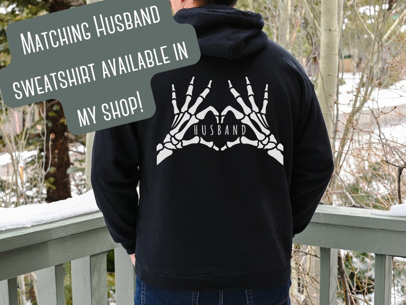 May include: Black hooded sweatshirt with a white skeleton hand design making a heart shape with the word "Husband" inside. The text "Matching Husband Sweatshirt Available in My Shop!" is superimposed on the image.