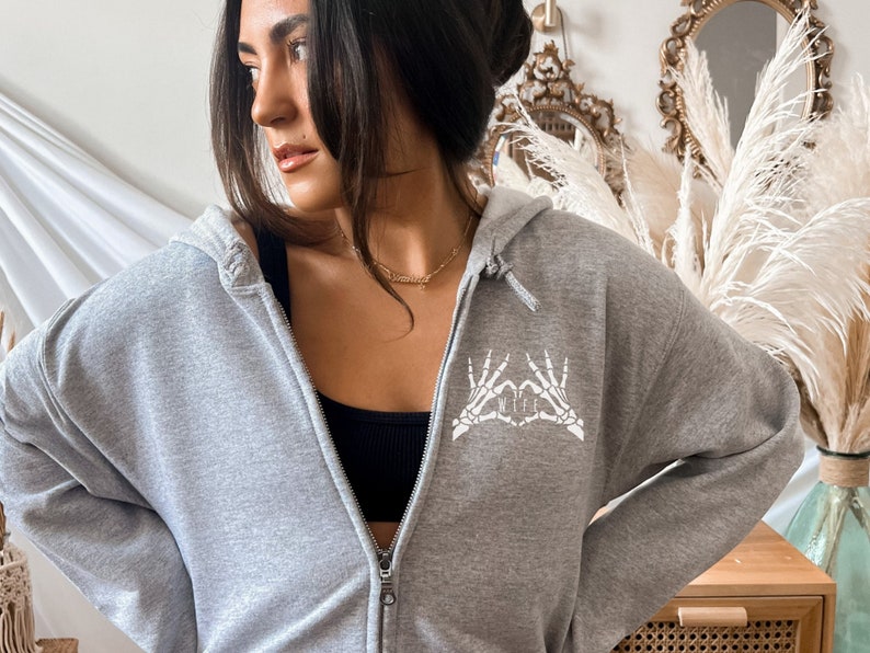 May include: Grey hooded sweatshirt with a white skeleton hand design that says "WIFE" in the centre of the heart.