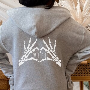 May include: Grey hooded sweatshirt with a white skeleton hand design making a heart shape with the word "WIFE" inside.