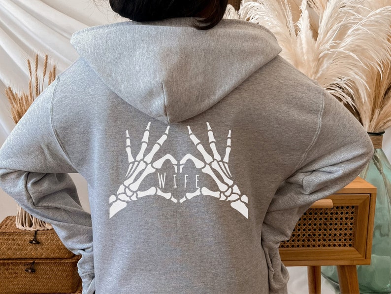 May include: Grey hooded sweatshirt with a white skeleton hand design making a heart shape with the word "WIFE" inside.