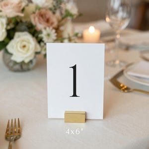 May include: A white rectangular table number card featuring the black printed numeral 1, held upright by a small gold-toned metal block stand. The card measures 4 by 6 inches and is displayed on a table setting with a floral centerpiece and cutlery.