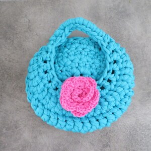 Könnte beinhalten: Eine kleine, runde, türkisfarbene Häkelhandtasche mit einem rosa Häkelblumen-Akzent.
