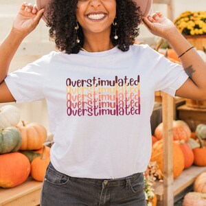 May include: A white t-shirt with the text "overstimulated" repeated in a gradient of orange, yellow, and red colors. The shirt is being worn by a person with brown skin and curly brown hair. The person is smiling and holding a wide-brimmed hat in their hands. The background is a wooden structure with pumpkins and fall foliage.