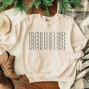 Op de afbeelding: Een beige sweatshirt met de tekst "HYPER FOCUSED" vijf keer herhaald in het zwart.
