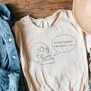 May include: A beige t-shirt with a black and white skull graphic and the text "My IBS? Chronic. This a**? Iconic."