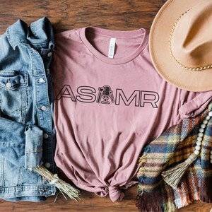 Op de afbeelding: Een mauve t-shirt met de tekst "ASMR" in zwarte omrande letters met een microfoonpictogram in het midden van het woord.