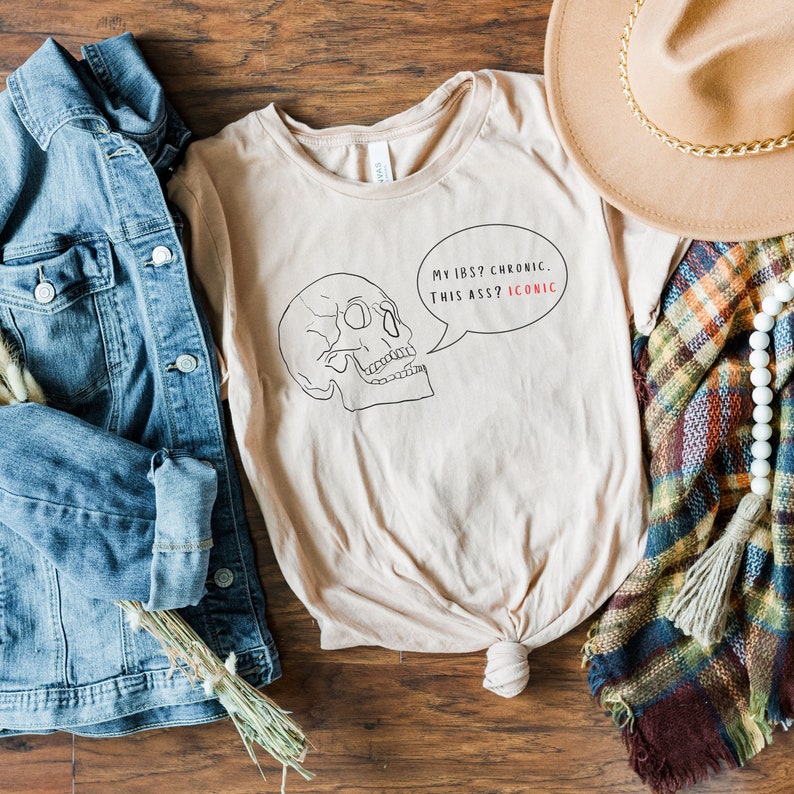 May include: A beige t-shirt with a black and white skull graphic and the text "My IBS? Chronic. This a**? Iconic."