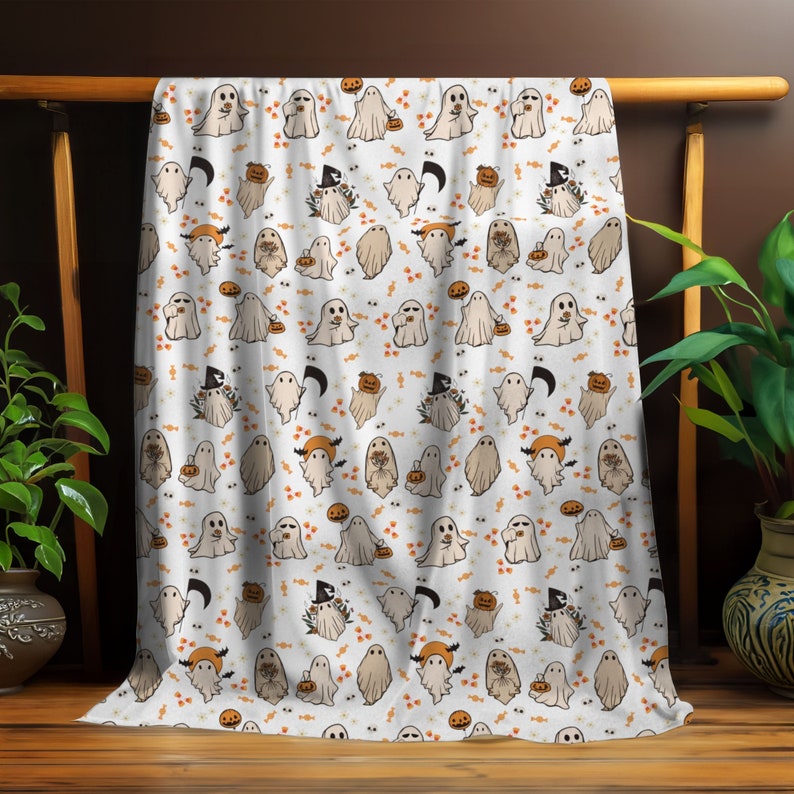May include: White throw blanket featuring a repeating pattern of adorable ghosts holding pumpkins and sweets. The ghosts are white with black eyes and mouths. The pumpkins are orange and the sweets are brown. The background is white with small orange and brown details.