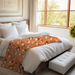 May include: Orange and white throw blanket with a Halloween pattern of ghosts, pumpkins, and bats. The blanket is on a white bed with white pillows.