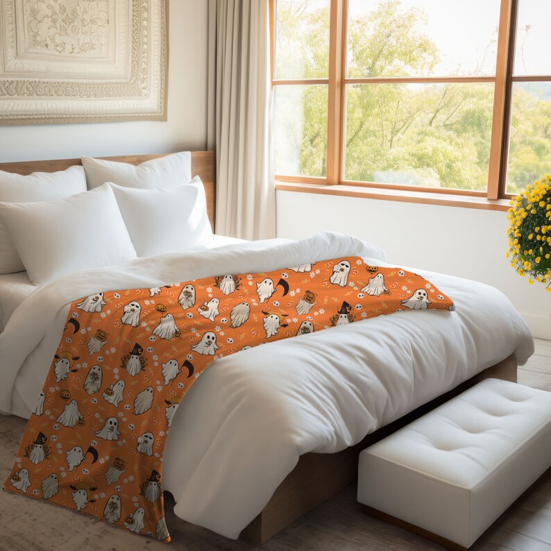 May include: Orange and white throw blanket with a Halloween pattern of ghosts, pumpkins, and bats. The blanket is on a white bed with white pillows.