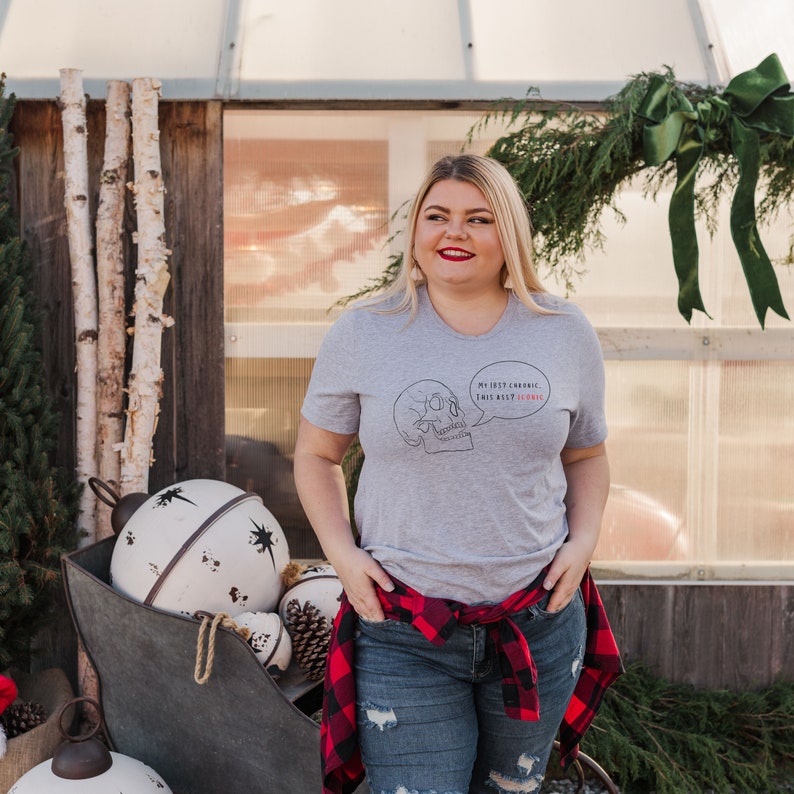 May include: A woman wearing a gray t-shirt with a skull graphic and the text "My life's chronic, this ain't a joke". She is wearing blue jeans with a red and black plaid shirt tied around her waist.