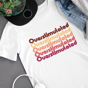 May include: White t-shirt with the text "Overstimulated" repeated five times in a retro font with orange, red, and yellow colors.