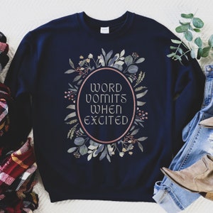May include: Navy blue sweatshirt with a floral wreath design and the text "WORD VOMITS WHEN EXCITED" in a white oval.