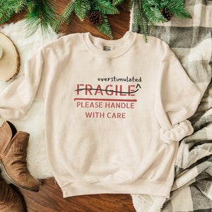 May include: A beige sweatshirt with the text "overstimulated FRAGILE PLEASE HANDLE WITH CARE" printed on the front. The word "FRAGILE" is crossed out and an arrow points up.