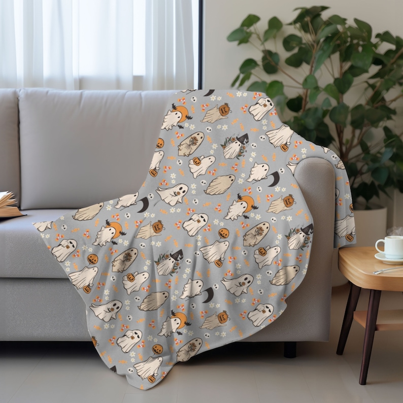 May include: A grey throw blanket with a repeating pattern of white ghosts holding pumpkins and candy corn. The ghosts are surrounded by small white flowers and skulls.