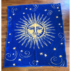 May include: A rectangular knit blanket featuring a vibrant blue background with a large, central yellow sun face. The sun is surrounded by numerous yellow stars and swirling line patterns scattered across the entire surface of the fabric.